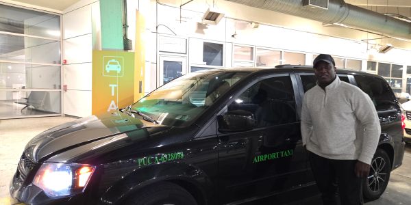 Man standing next to a black airport taxi vehicle inside a parking area.