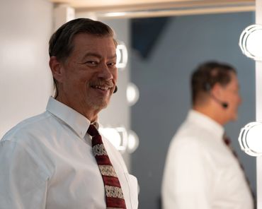 Smiling man in a white shirt and patterned tie with headset, standing near a lighted mirror.