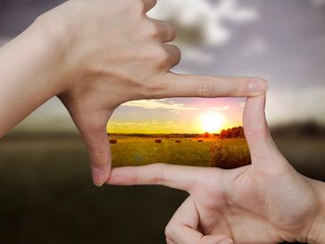 Hands framing a vibrant sunset over a hayfield.
