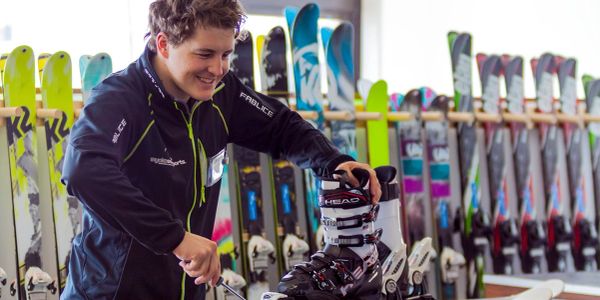 A person adjusting a ski boot on a rack with colorful skis in the background.