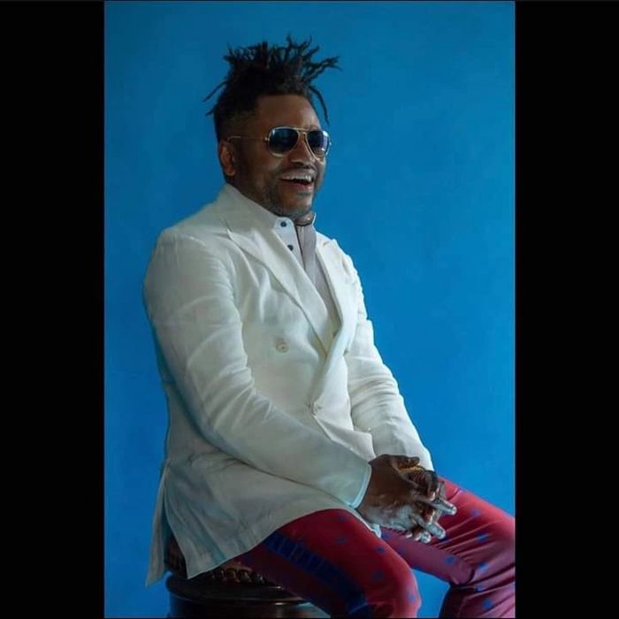 Rakiem Walker, musician, posing, smiling sitting on stool; wearing white suit jacket and red pants.