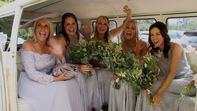 Five joyful bridesmaids in matching dresses waving inside a vehicle with bouquets.