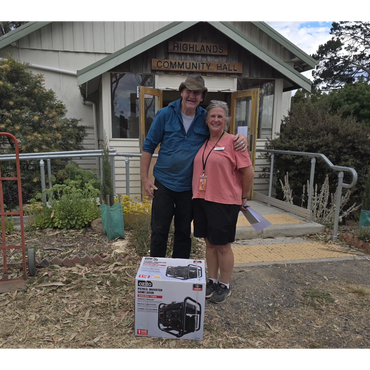 Two people smiling in front of Highlands Community Hall with a boxed generator.