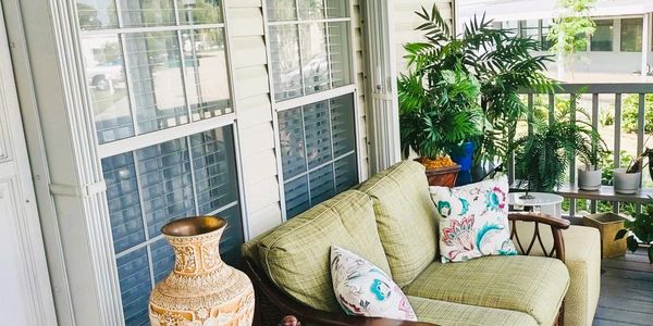 Cozy porch with green sofa, decorative pillows, large vase, and lush plants.