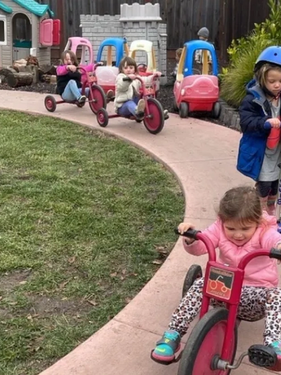 Children riding tricycles and scooters on a curved path in a playground.