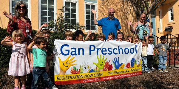 Children and teachers happily waving behind a Gan Preschool registration banner.