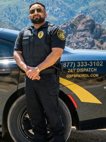 A security officer stands confidently by a patrol car in a mountainous area.