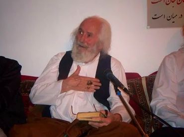 Elderly man speaking while holding a book and gesturing with his hand.