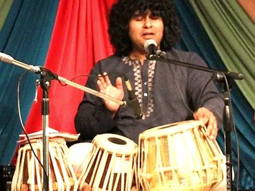 Musician playing tabla and singing on stage with colorful backdrop.