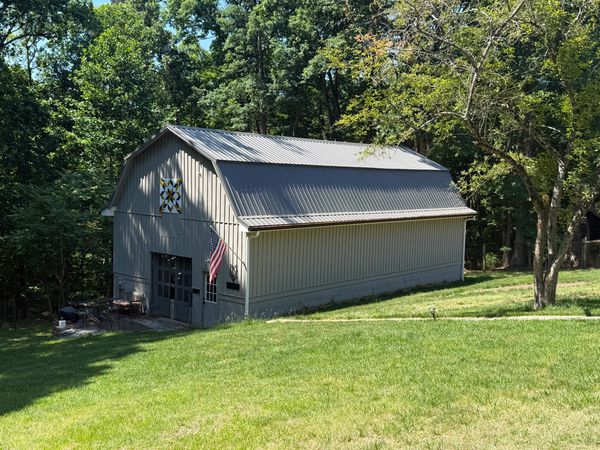 A gray barn with an American flag and a quilt pattern in a green wooded area.
