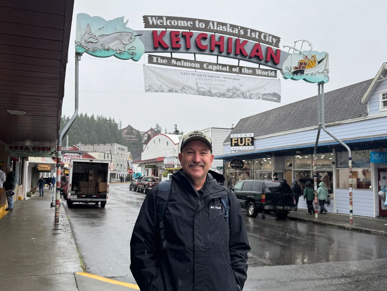 Man stands smiling under Ketchikan welcome sign on rainy street.