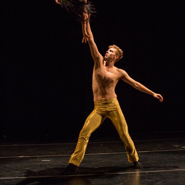 Male dancer in gold pants reaching upwards on stage.