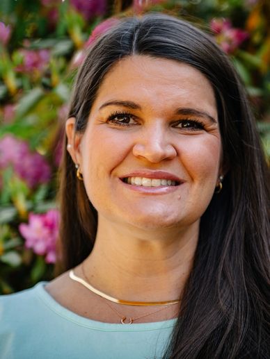 Picture of a woman with dark hair standing outside in front of pink flowers on a bush