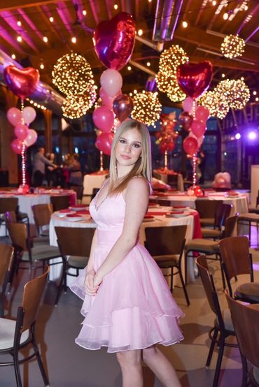Young woman in a pink dress at a decorated party with heart-shaped balloons and lights.