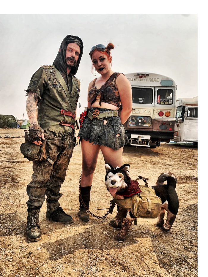 A wasteland couple in tattered costumes and boots with a stuffed Dogmeat dog in front of a schoolbus