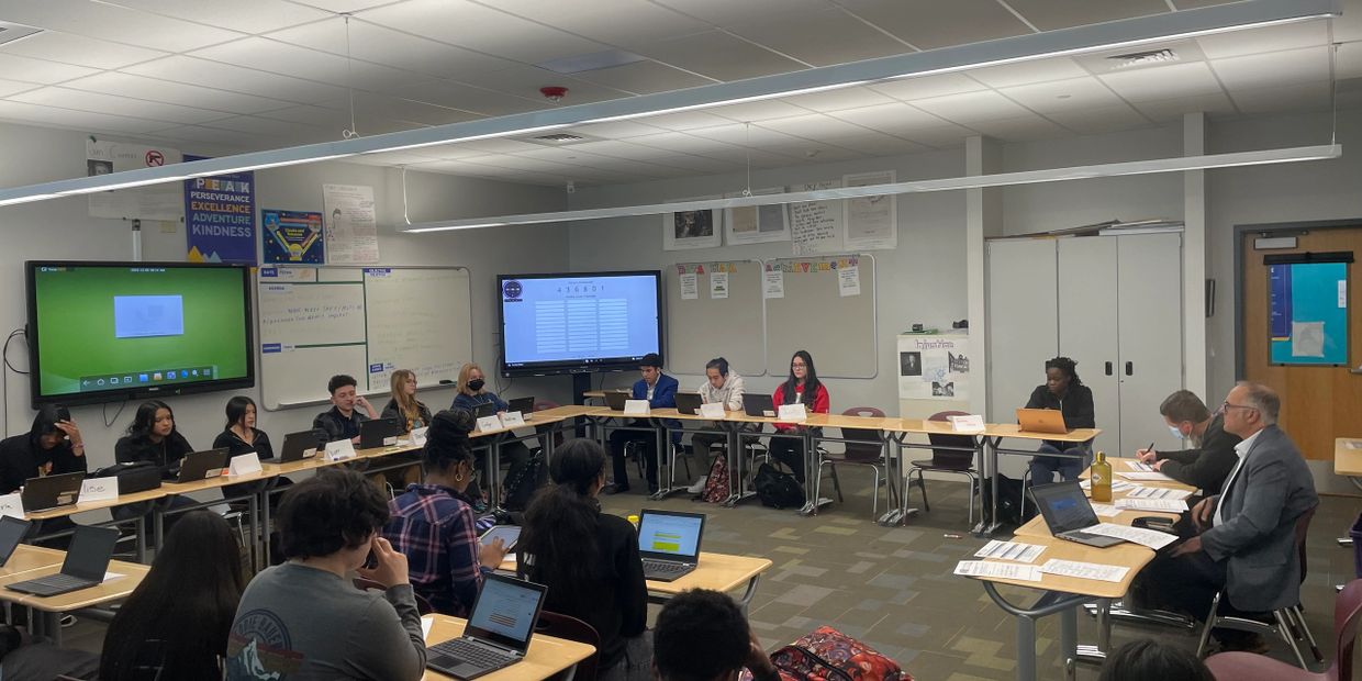 Students and teachers engaged in a classroom discussion with laptops and screens.