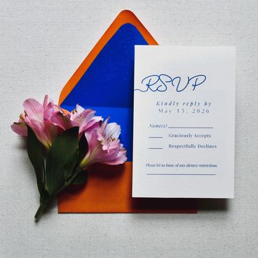 RSVP card with pink flowers and a vibrant orange and blue envelope on a textured surface.