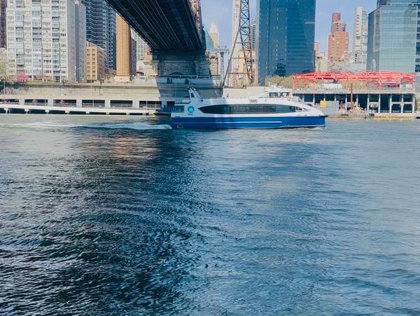 A ferry boat passes under a large city bridge with skyscrapers in the background.