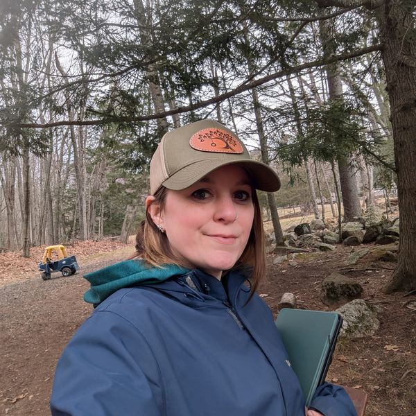 Woman in outdoor jacket and cap in a wooded area, holding a tablet.
