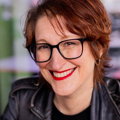 Julie Cohen smiles at the camera. She has black-framed glasses on, short hair and a leather jacket.