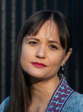 Photo of Natalie Gregory, with a corrugated metal background and a breeze blowing stands of hair.