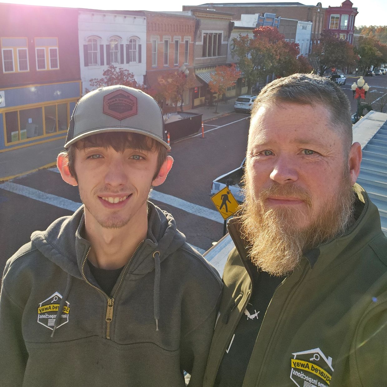 Two men wearing jackets and a cap, smiling outdoors in a small town setting.