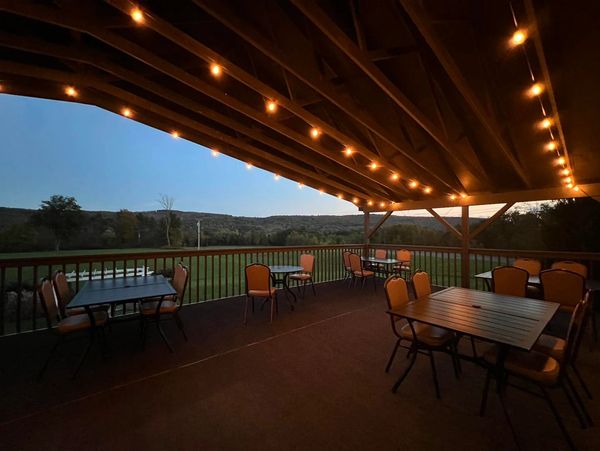 Outdoor covered patio with string lights and seating, overlooking a green field and hills at dusk.