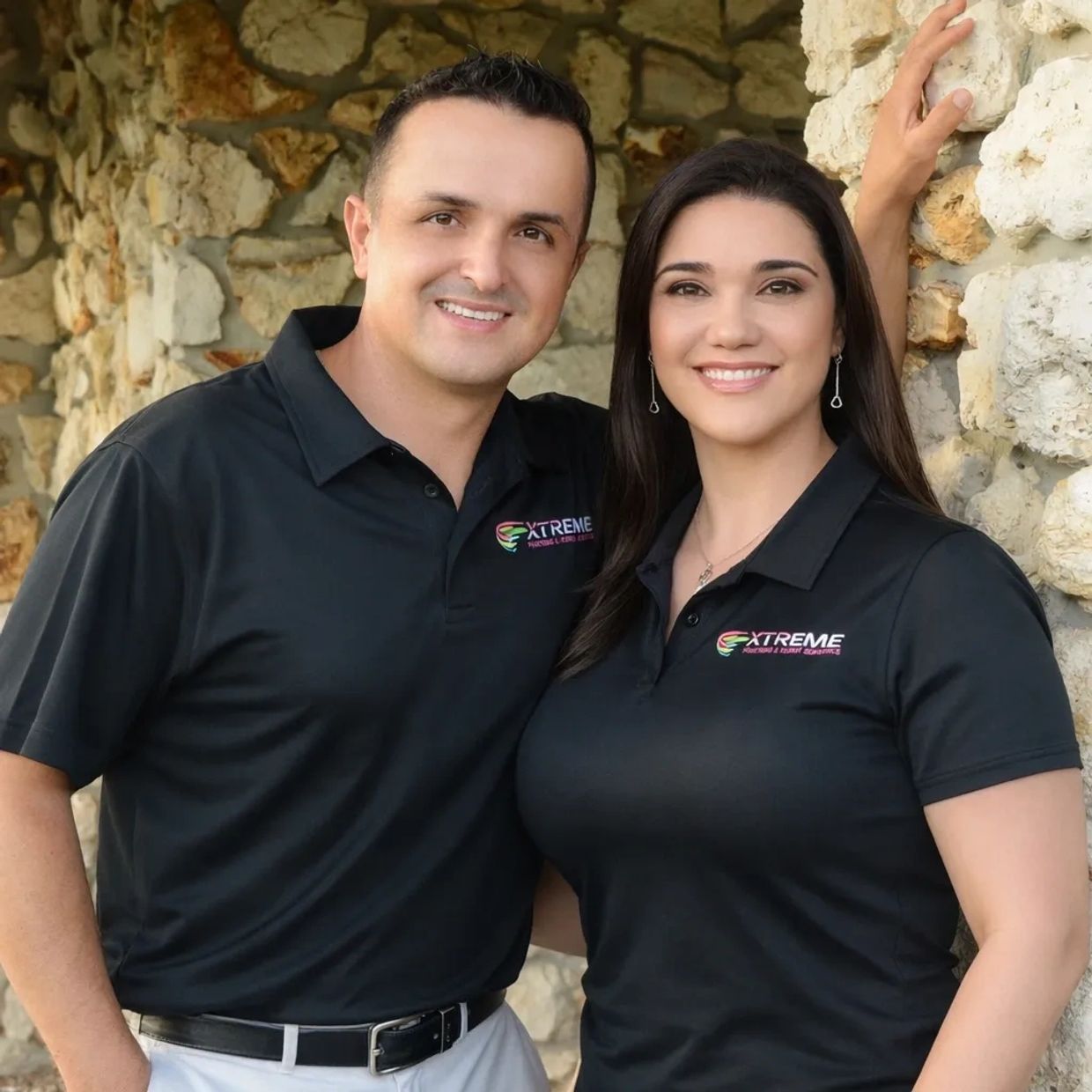 Smiling man and woman in black company polo shirts standing against a stone wall.