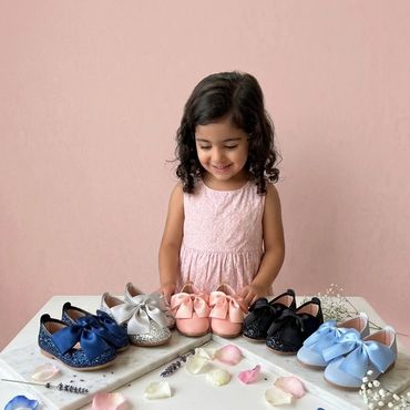 Smiling girl admiring colorful bow-adorned shoes on a table with scattered flower petals.
