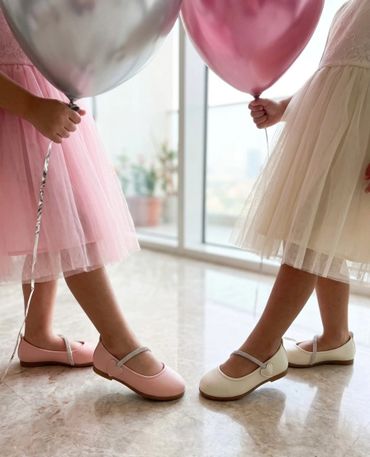 Two children in pastel dresses holding balloons and wearing matching Mary Jane shoes.