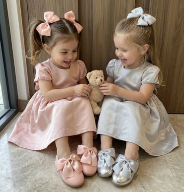 Two young girls in pastel dresses sharing a teddy bear and smiling.
