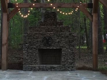 Stone outdoor fireplace decorated with string lights in a wooded area.