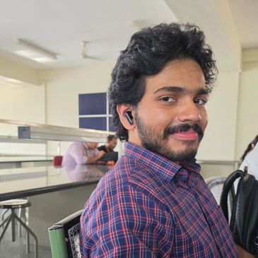 Man with curly hair and beard wearing a checkered shirt and earphones indoors.
