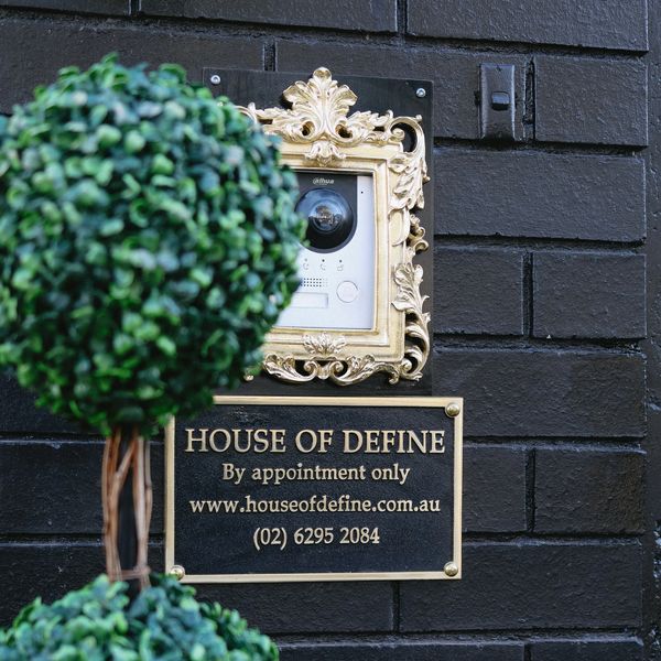 Security camera framed ornately above a sign for House of Define on a black brick wall.