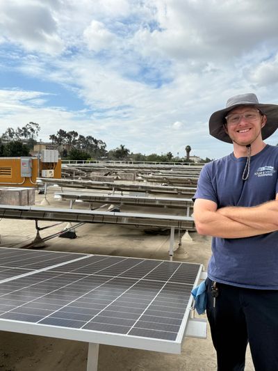 A man stands smiling next to solar panels under a cloudy sky.