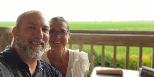 Smiling couple enjoying a bright day on a wooden deck overlooking green fields.