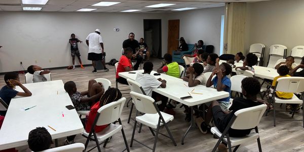 Children seated around tables in a classroom setting, listening to an instructor.