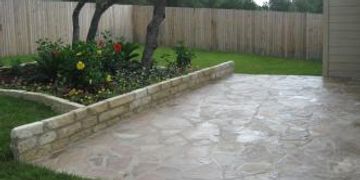 Stone patio with a raised flower bed bordered by a low stone wall in a backyard.