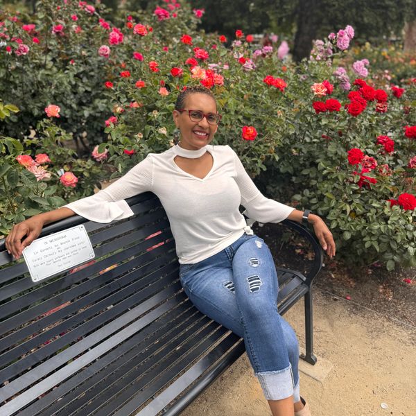 Woman in white top and jeans relaxing on a bench surrounded by colorful roses.