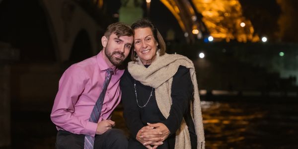A smiling couple posing by a riverside at night with warm lights in the background.