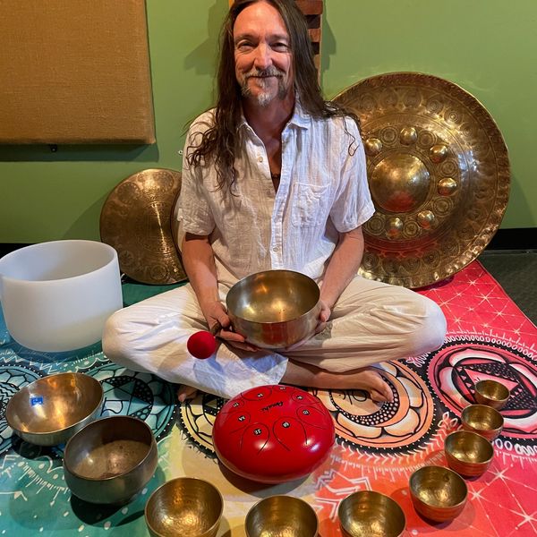a joyful man sitting with many tibetan sound bowls, a red hand pan drum and a chinese wind gong