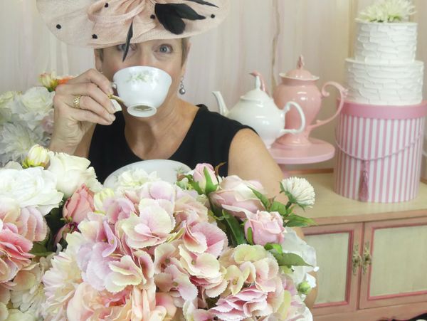 Woman wearing a fascinator and drinking tea....surrounded by flowers and a tea pot.