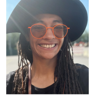 Person wearing large hat and orange glasses smiling.
