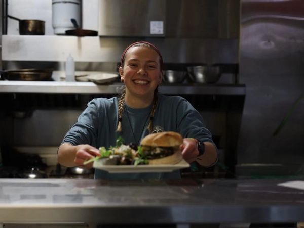 Smiling chef holding a plate with a sandwich and salad in a stainless steel kitchen.