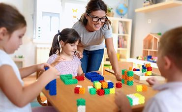 A teacher helps children build colorful block structures at a table.