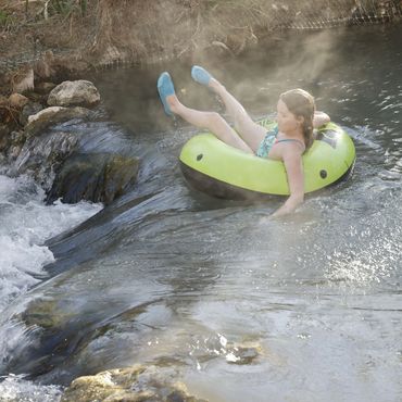 A girl in a green tube floats on steaming water near a small waterfall.