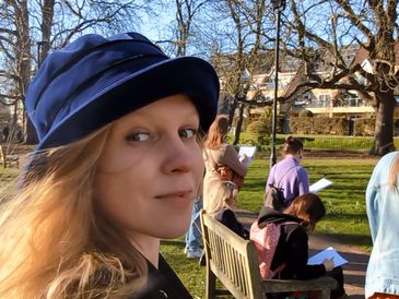 Woman in blue hat smiling outdoors with others reading in park.