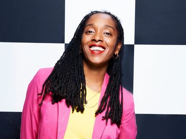 A smiling woman with long dreadlocks wearing a pink blazer and yellow shirt.