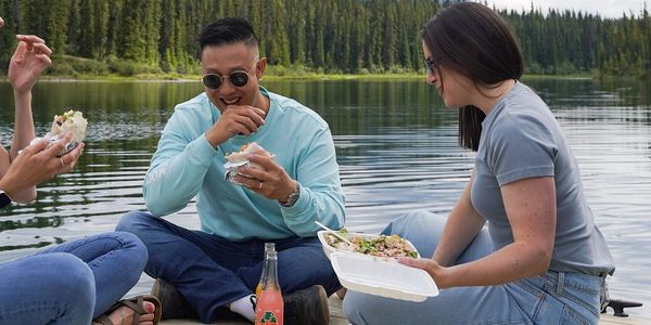 Three friends eating by a lake with trees and mountains in the background.