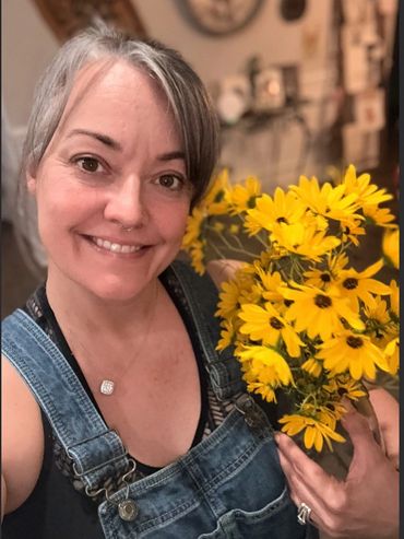 Smiling woman in denim overalls holding bright yellow flowers.